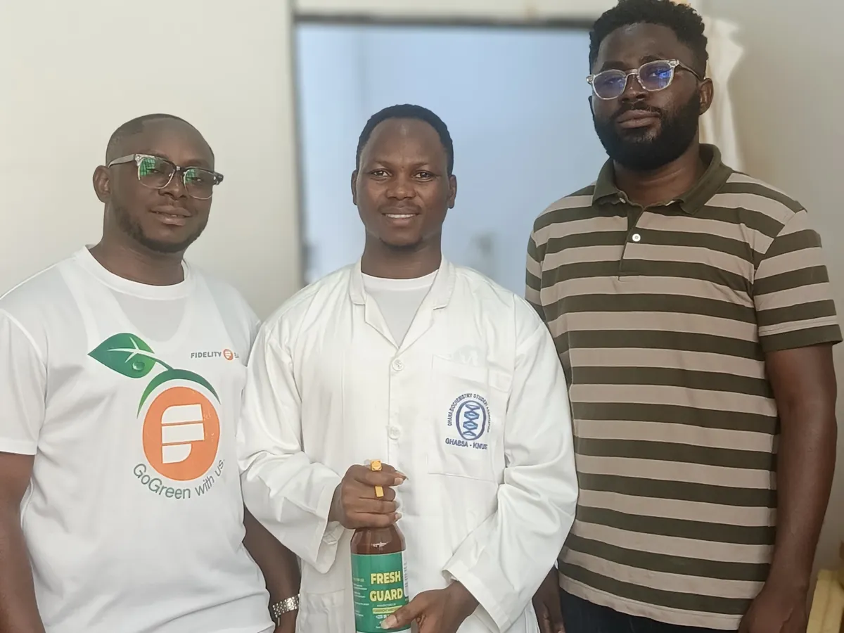 A researcher in a lab coat holding a FreshGuard bottle, standing between two visitors, one in a Fidelity Bank GoGreen T-shirt